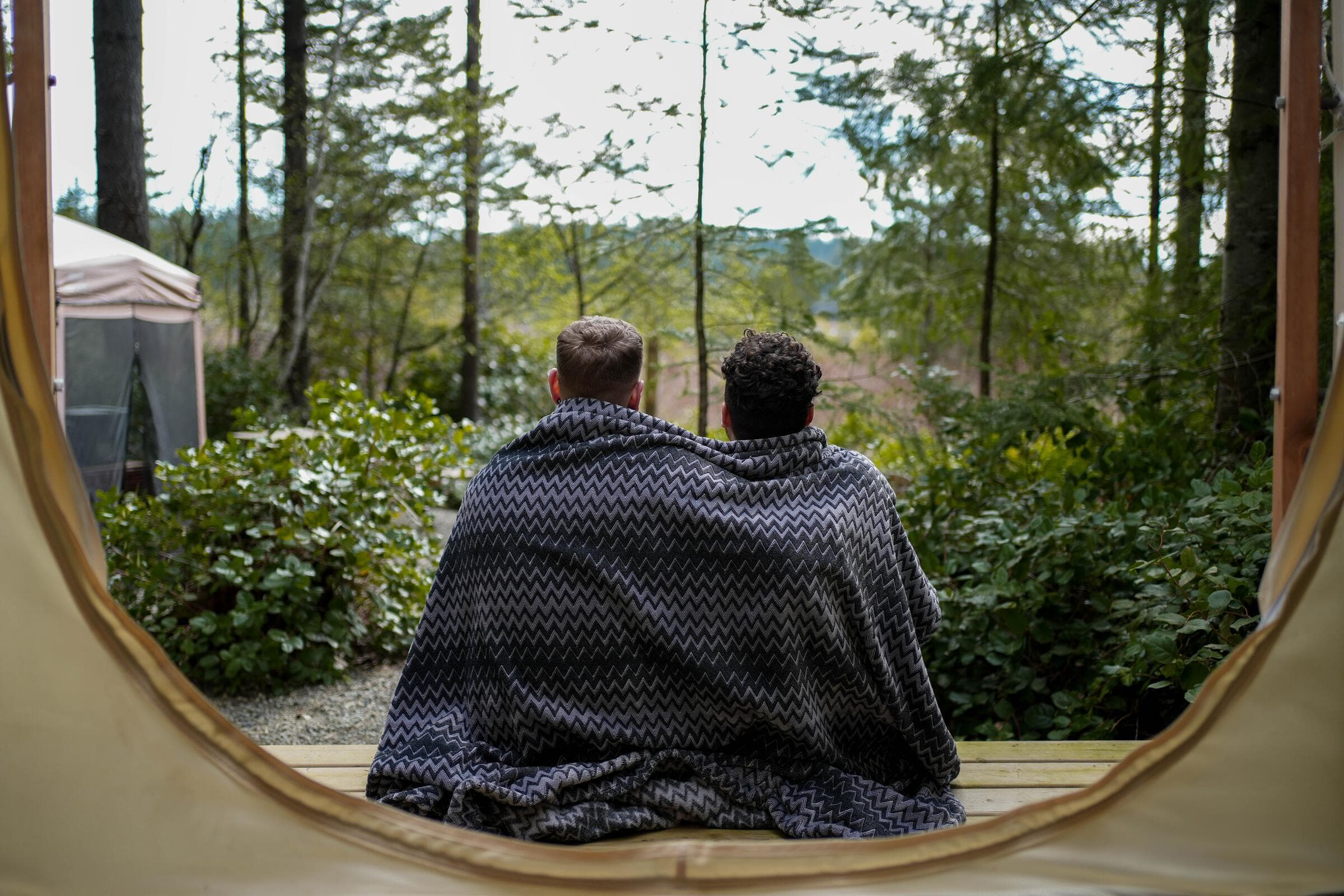 Two people wrapped in a blanket on the deck of a glamping tent in the forest near Merville