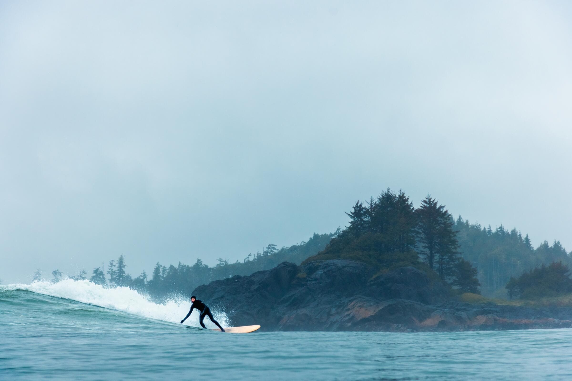 A surfer on a small wave beside a rocky, tree-covered islet near Tofino